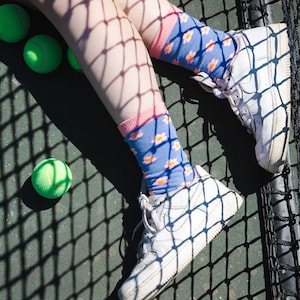 Könnte beinhalten: Ein Paar weiße Sneaker mit blauen und rosa Socken mit Blumenmuster. Die Sneaker befinden sich auf einem grünen Tennisplatz mit einem schwarzen Netz.