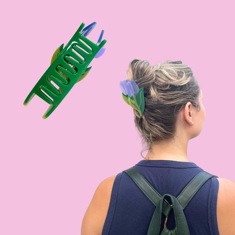 K&ouml;nnte beinhalten: Eine gr&uuml;ne Haarspange in Tulpenform mit violetten Bl&uuml;tenbl&auml;ttern und gr&uuml;nen Bl&auml;ttern. Die Spange verf&uuml;gt &uuml;ber einen Federmechanismus mit ineinandergreifenden Z&auml;hnen, um das Haar zu halten. Das Design zeigt einen gr&uuml;nen Stiel und Bl&auml;tter.