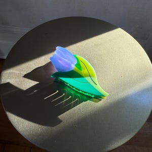 K&ouml;nnte beinhalten: Eine Haarspange in Form einer Tulpenbl&uuml;te. Der obere Teil ist durchscheinend hellviolett, w&auml;hrend die Basis und die blattartige Struktur in einem leuchtenden, halbtransparenten Gr&uuml;n gehalten sind, akzentuiert durch feine dunkelgr&uuml;ne Linien.