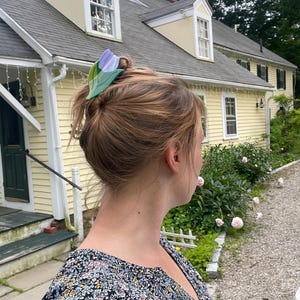 K&ouml;nnte beinhalten: Eine Haarspange in Form einer Tulpenbl&uuml;te mit einem hellvioletten Bl&uuml;tenblatt und einer gr&uuml;nen Basis aus Stiel und Blatt. Die Spange ist in hellbraunem Haar befestigt und h&auml;lt einen Teil des Haares in einem l&auml;ssigen Stil zusammen.