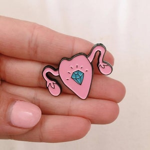 Könnte beinhalten: Rosa emaillierte Anstecknadel in Form einer Gebärmutter mit einem blauen Diamanten in der Mitte. Die Nadel hat einen schwarzen Umriss.