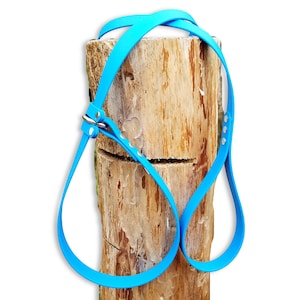 May include: A bright cyan pet harness featuring a smooth, flexible strap design with silver-toned metal hardware, including a buckle and adjustment holes, draped over a textured, light brown cylindrical wooden log display piece.