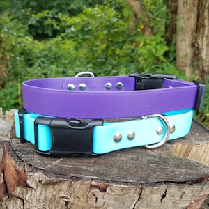 May include: Two dog collars, one purple and one light blue, with silver metal accents and black buckles. The collars are stacked on top of a tree stump.