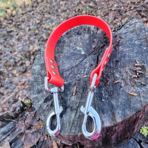 May include: A red dog leash with two silver clips. The leash is made of a durable, rubber-like material and is lying on a wooden surface.