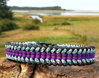 Puede incluir: Una pulsera de paracord con un diseño trenzado de varias capas en negro, morado vibrante y un patrón moteado en azul claro y blanco. La pulsera se asegura con una hebilla de liberación lateral de plástico negro, mostrando una textura trenzada.