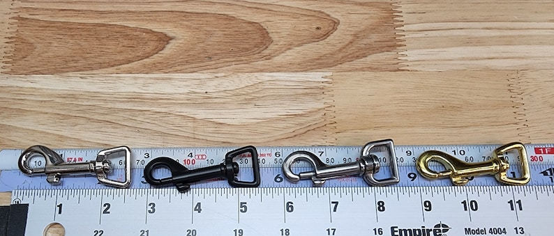 May include: Five metal swivel snap hooks in different finishes: silver, black, nickel, and gold. The hooks are lined up on a ruler with measurements in inches.