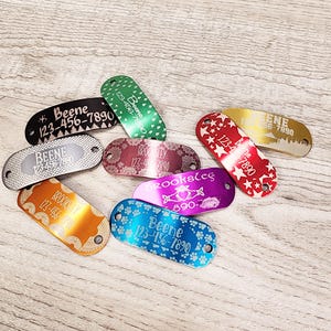 May include: A selection of colourful metal dog tags with engraved names and phone numbers. The tags feature a variety of designs, including paw prints, stars and Celtic knots.