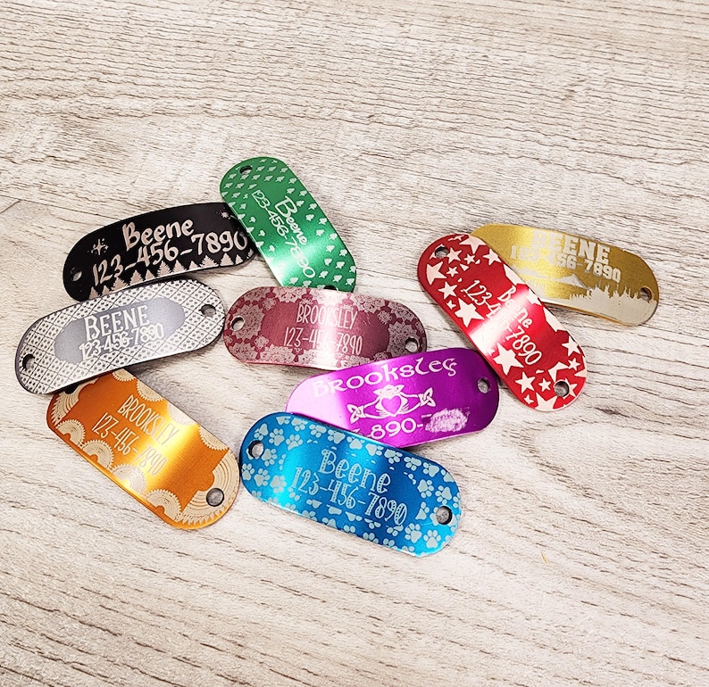 May include: A selection of colourful metal dog tags with engraved names and phone numbers. The tags feature a variety of designs, including paw prints, stars and Celtic knots.