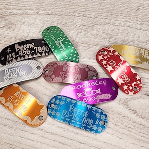 May include: Assortment of colourful metal dog tags with engraved names and phone numbers. The tags feature various designs including stars, paw prints, and trees.