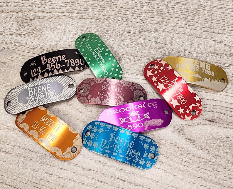 May include: Assortment of colourful metal dog tags with engraved names and phone numbers. The tags feature various designs including stars, paw prints, and trees.