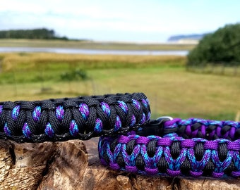 May include: Two braided paracord collars featuring a black, purple, and blue woven pattern. Each collar includes a metal D-ring attachment point and a black plastic side-release buckle, showcasing a thick, textured design made from durable cord.