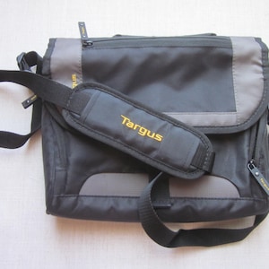 May include: A black and gray Targus messenger bag featuring a front flap, multiple zippered compartments with branded zipper pulls, and an adjustable shoulder strap with a padded shoulder rest displaying the yellow Targus logo in embroidery.