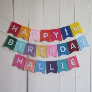 Puede incluir: Una colorida guirnalda de fieltro con las palabras "HAPPY 1st BIRTHDAY HALLIE" en letras blancas. La guirnalda presenta banderas de varios colores, incluyendo rojo, amarillo, rosa, verde y azul, unidas por una cuerda blanca.
