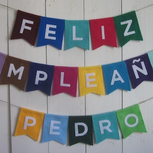 May include: A colorful felt banner with the words "Feliz Cumpleaños Pedro" in white letters. The banner is strung on a white string and is hanging against a white wooden background.