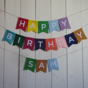 May include: A colorful felt banner with the words "Happy Birthday Sam" in white letters. The banner is strung on a white wall.
