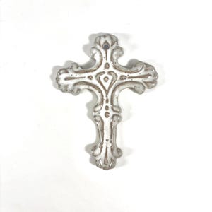 May include: A white ceramic cross ornament featuring a glossy finish with brown speckled accents and intricate scrollwork detailing. The cross has a small circular hole at the top for hanging and displays a symmetrical, ornate shape with curved edges.