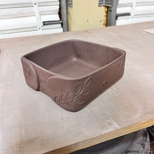 May include: A square, brown ceramic dish with a leaf design on the side. The dish is sitting on a wooden surface.