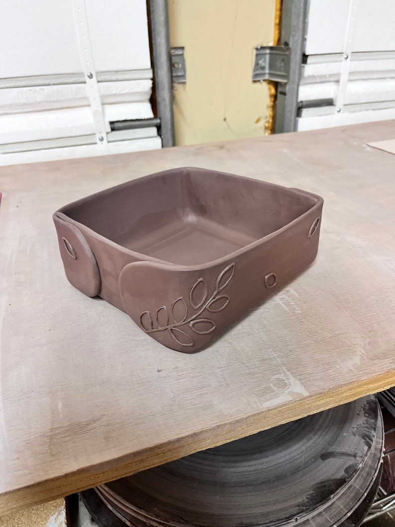 May include: A square, brown ceramic dish with a leaf design on the side. The dish is sitting on a wooden surface.