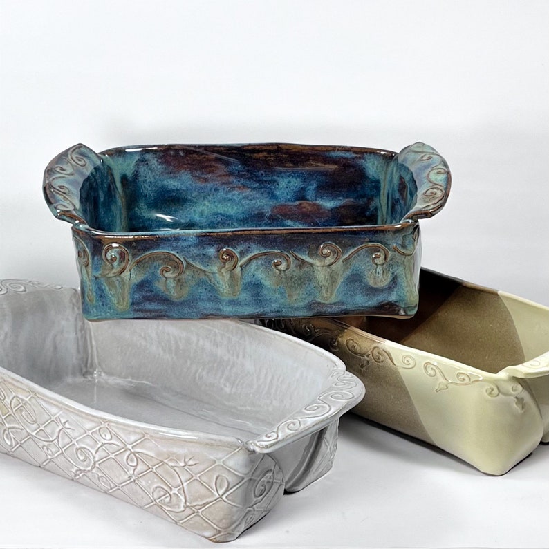 May include: Three ceramic baking dishes with decorative edges. The top dish is teal blue with brown accents. The middle dish is white with a subtle embossed pattern. The bottom dish is beige and brown with a swirl pattern.