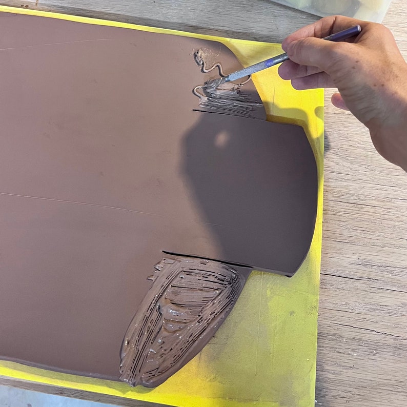 May include: A person is using a brush to apply brown paint to a clay surface. The clay is on a yellow surface.