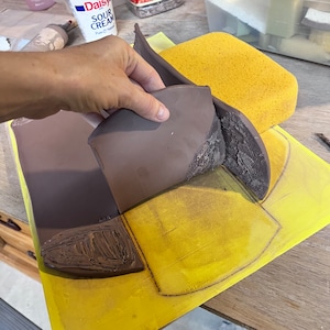 May include: A brown clay pot being shaped on a yellow surface with a pencil outline. The pot is being molded by hand.