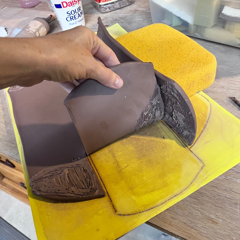 May include: A brown clay pot being shaped on a yellow surface with a pencil outline. The pot is being molded by hand.