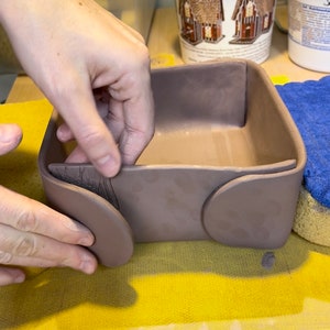 May include: A person is attaching a brown clay wheel to a brown clay box. The box has a square base and four sides. The wheel is round and has a flat surface.