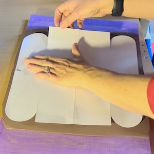 May include: A white paper template is being placed on a brown clay surface. The template is divided into four sections, each with a curved edge. The clay surface is surrounded by a purple mat.