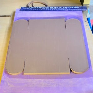 May include: A light brown clay tile with two rounded cutouts on each side. The tile is sitting on a purple surface.