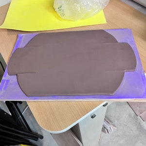 May include: A brown clay slab with a rounded shape and a cut-out design, resting on a purple mat.