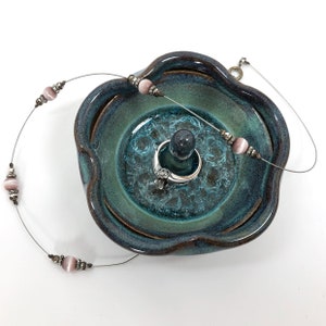 May include: A teal and brown ceramic jewelry dish with a central ring holder. A silver ring sits on the holder. A delicate necklace with pink beads and silver accents rests on the dish. The dish has a wavy edge.