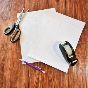 May include: Two white paper sheets with a heart shape drawn on each sheet. A purple pencil, a pair of silver scissors, and a black tape dispenser are on a wood surface.