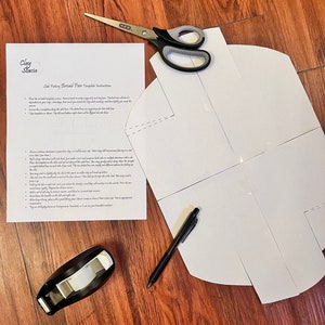 May include: A white paper template for a bread pan, with instructions on how to fold it. The template is on a wooden surface with a pair of scissors, a pen, a tape dispenser, and a roll of tape.