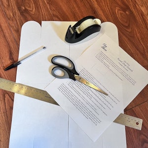 May include: A white paper template for a round strap casserole with instructions on how to assemble it. The template is surrounded by a ruler, scissors, tape dispenser, and a pen.