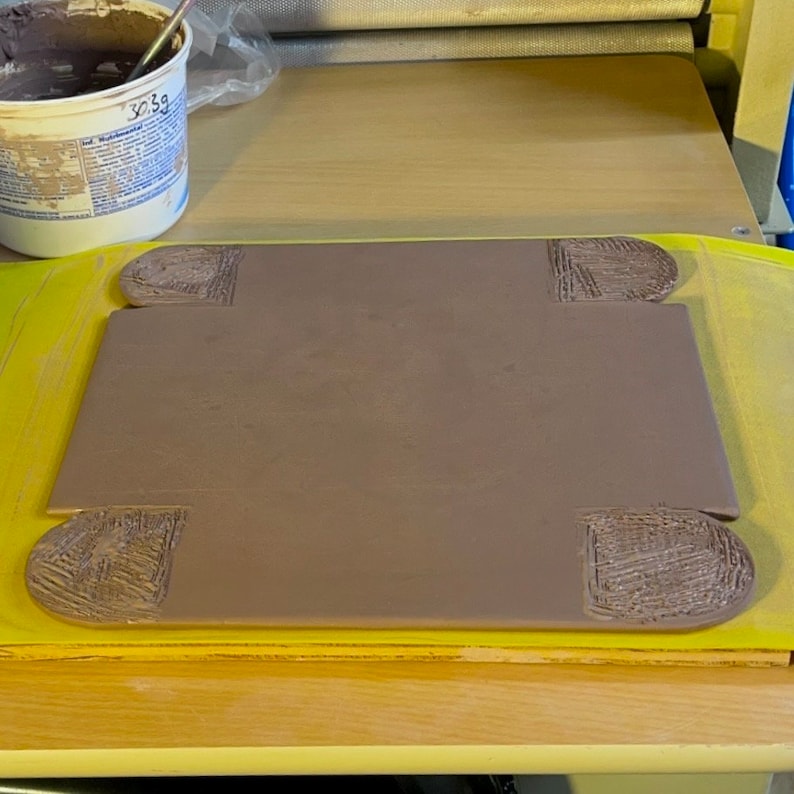 May include: A brown clay tile with four raised edges, sitting on a yellow surface. The tile has a textured surface and is ready for crafting.