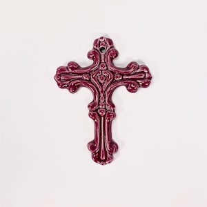 May include: A burgundy-colored decorative cross with ornate detailing. The cross features a heart-shaped design in the center and decorative flourishes on the arms and base. The cross is made of a glossy material and has a hole at the top for hanging.