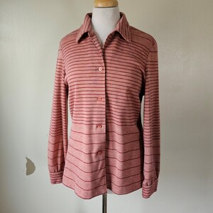 May include: A button-down shirt with long sleeves, displayed on a mannequin. The shirt is a light reddish-pink color with horizontal stripes in a darker reddish-brown. The shirt has a collared neckline and is buttoned up the front.