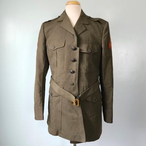 Puede incluir: Chaqueta militar verde oliva con cinturón en la cintura y cuatro bolsillos delanteros. La chaqueta tiene cierre de botones y charreteras en los hombros. Un pequeño parche es visible en la parte superior del brazo. La chaqueta se exhibe en un maniquí.