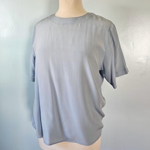 May include: A short-sleeved blouse in a solid light blue color. The garment features a simple round neckline and a smooth, slightly lustrous fabric finish. The cut is relaxed with a straight hemline, providing a clean and straightforward appearance.