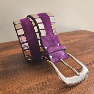 Op de afbeelding: Een paarse suède riem met zilveren vierkante studs en een zilveren gesp. De riem wordt getoond op een houten oppervlak, waarbij de textuur en het ontwerp worden benadrukt. De gesp van de riem heeft een grote, zilveren, rechthoekige vorm.