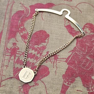 May include: A gold-toned tie bar featuring a curved, arched top bar connected by two delicate curb chains to a central, octagonal pendant. The pendant is engraved with the capital letter B in a serif font, creating a classic, structured accessory.