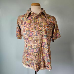 Op de afbeelding: Een shirt met korte mouwen met een geometrisch patroon in geel, roze, paars en bruin. Het shirt wordt op een mannequin getoond en heeft een kraag en een knoopsluiting.