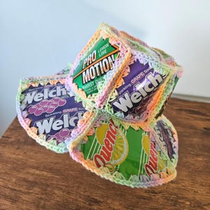 May include: A stack of colorful, crocheted drink can cozies. The cans feature the Welch's logo and the words "Sparkling Grape Soda". Other cans say "Pro Motion" and "Quench". The crochet work is in pastel colors.