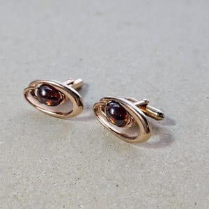 May include: A pair of gold-coloured cufflinks. Each cufflink has a dark amber-coloured, oval-shaped stone set within a gold oval frame. The cufflinks have a classic, elegant design.