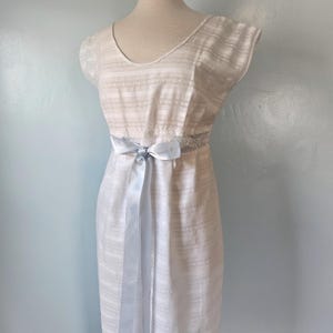 May include: A vintage-style, cream-colored dress with a subtle striped pattern. The dress features a scoop neckline, cap sleeves, and a light blue satin ribbon tied at the waist. The dress is displayed on a mannequin.