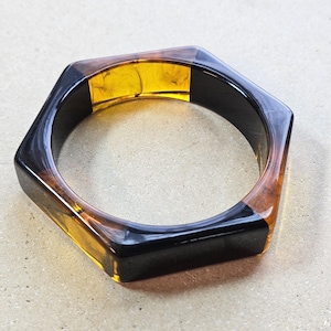 May include: Hexagonal bangle bracelet with a geometric design. The bracelet features a combination of black, amber, and brown colors. The bracelet is made of a glossy material.