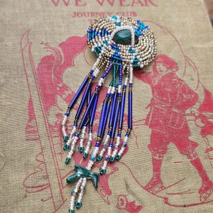 May include: A circular beaded brooch featuring a central dark green cabochon surrounded by concentric rings of metallic silver, blue, and green seed beads. Long, dangling fringe strands of blue, white, and green beads hang from the base.