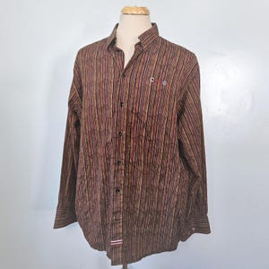 Puede incluir: Una camisa de manga larga con botones, de color base marrón con rayas verticales en rojo, amarillo y negro. La tela tiene una apariencia texturizada y arrugada. El texto "CDG" está bordado en el pecho izquierdo y una pequeña etiqueta a rayas rojas y blancas es visible en el dobladillo inferior.