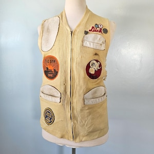 May include: A pale yellow sleeveless vest featuring a front zipper, multiple white pockets, and various embroidered patches including one that reads "Tucson 1968", "Dick", "RMD", and "ATA Life Member". The vest shows signs of wear with small holes.