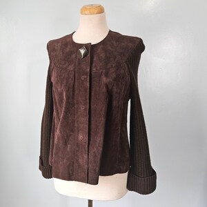 May include: A brown suede jacket with a unique design. The jacket features a button-up front, a decorative silver clasp, and knit sleeves. The jacket is displayed on a mannequin against a light gray background. The jacket's sleeves have a ribbed cuff.